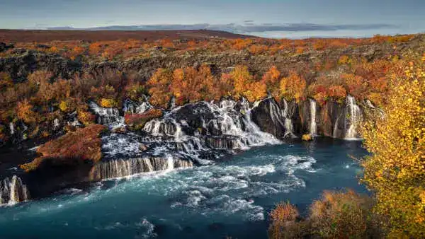 Multiple cascades qui coulent d'un champ de lave vers une rivière bleue turquoise, avec les couleurs de l'automne