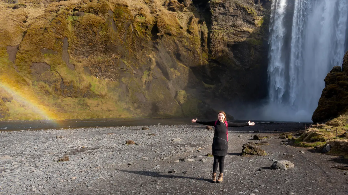 Une femme se tient les bras ouverts devant une cascade et un arc-en-ciel