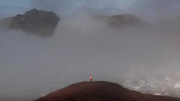 A man in an orange jacket is on top of a hill overlooking a town further down