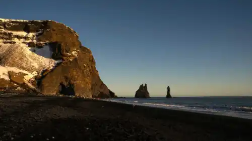 A black sand beach, with a cave and sea pillars is photographed at sunset