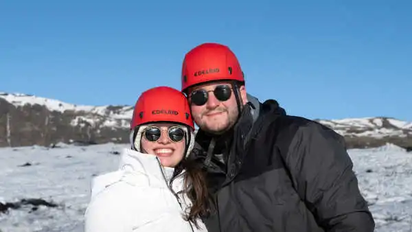 A couple is smiling in front of the camera, on a glacier, they wear a red helmet