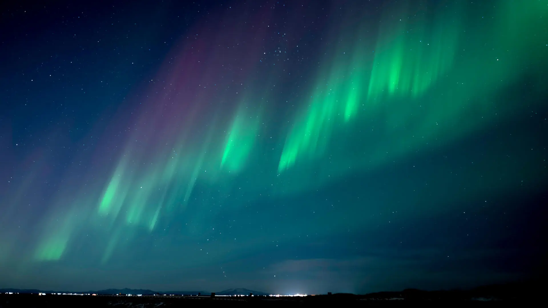 Green and purple Northern Lights are shining over a glacier and a small town