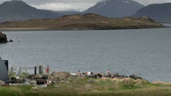 People are bathing in hot springs by the ocean, surrounded by mountains