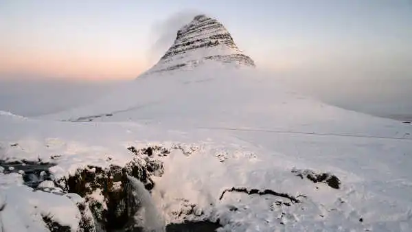 Pink skies during sunrise, with a snow covered, pointy mountain in the background and two waterfalls in the front