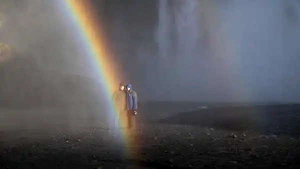 Two people are standing in front of a huge waterfall, and are touched by a rainbow