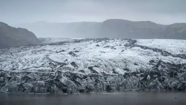 The end of a white and blue, ash covered, glacier is touching the water in a misty day