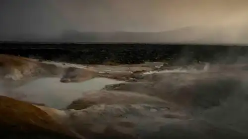 Hot springs steaming from everywhere are creating a fog in front of colourful moutains, under a soft, warm, sunset light.